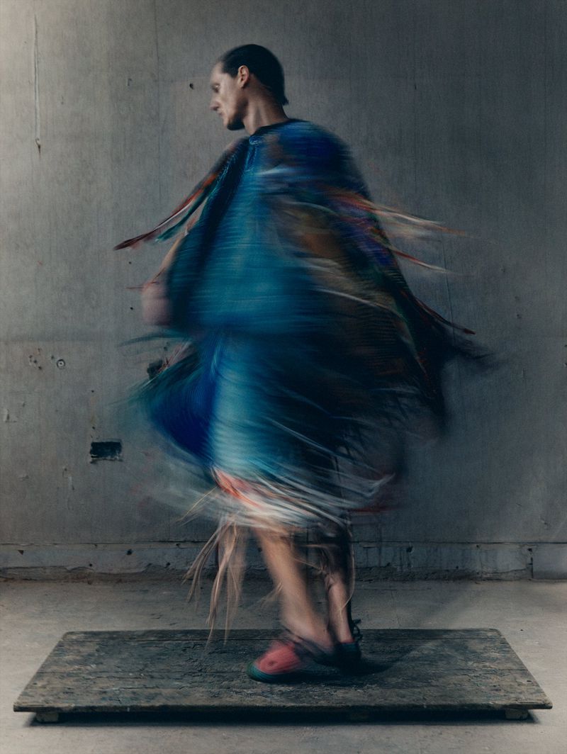 Adult male in motion wearing fringed blue garment and red shoes on a platform against a concrete wall, motion blur, soft light.