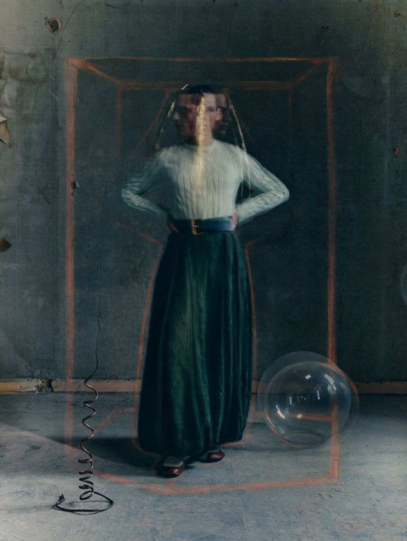 Woman in sweater and green skirt standing in old room with orange frame. Portrait-style fashion shot in desaturated color palette.