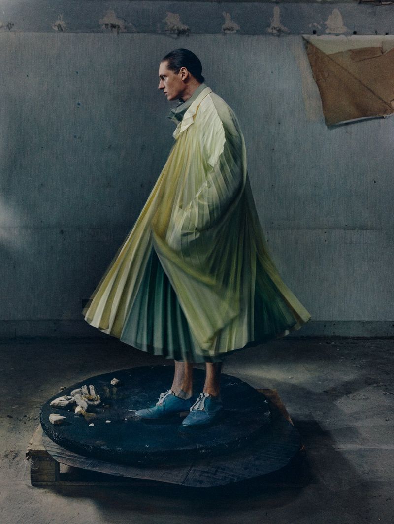 A person in a long yellow coat and blue shoes stands on a black platform in a photo studio with neutral wall background, fashion shot.