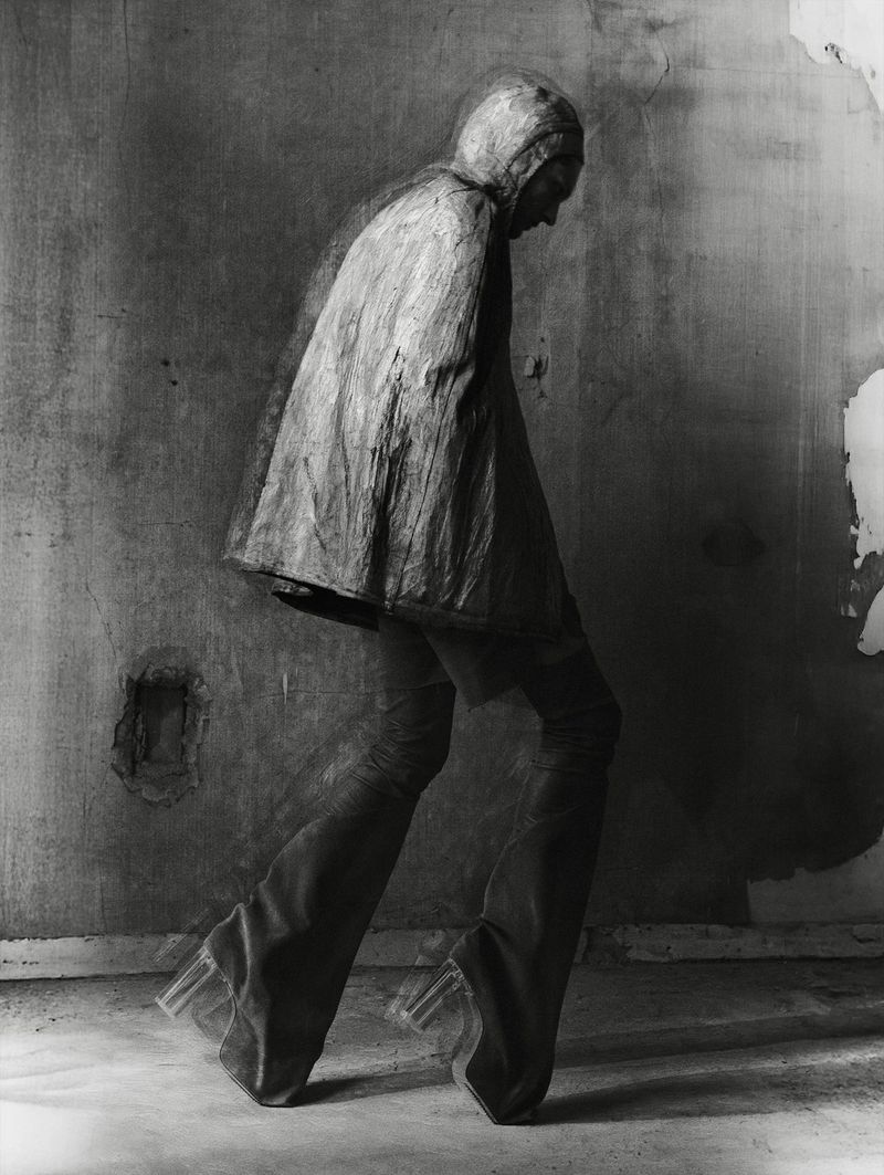 Monochrome fashion shot of a person in a hooded garment, pants, and block heel shoes against a distressed wall, walking forward.