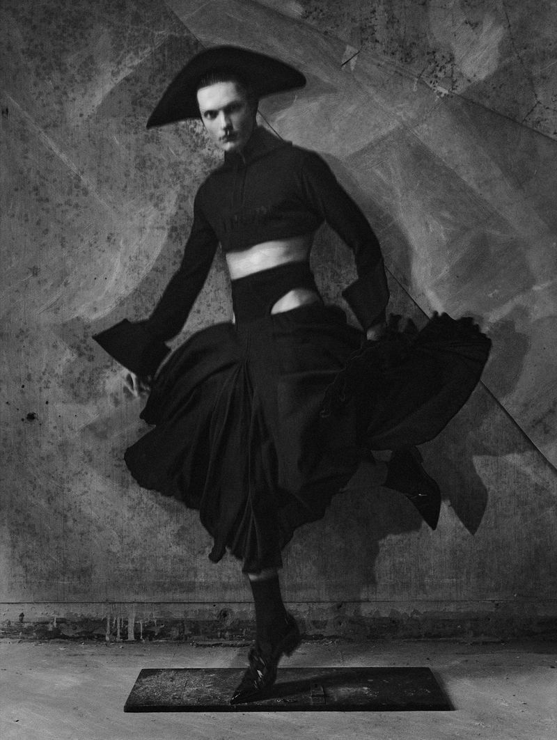 Elegant portrait of a dancer in avant-garde black fashion attire, featuring a wide-brimmed hat, crop top, and voluminous skirt in a studio setting.