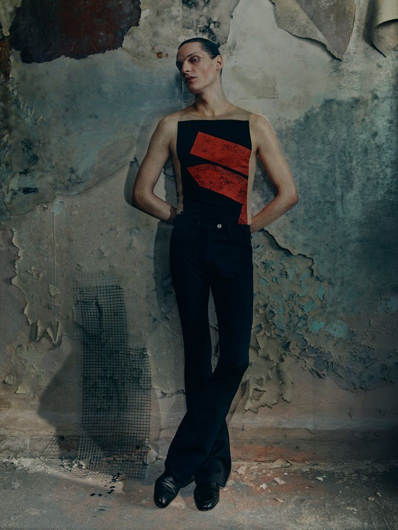 An adult male fashion model poses against a distressed interior wall wearing a black outfit with red accents and leather shoes.