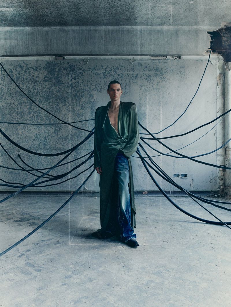 Adult male stands in a decayed interior wearing a robe and jeans, surrounded by black cables against a textured grey wall and floor.