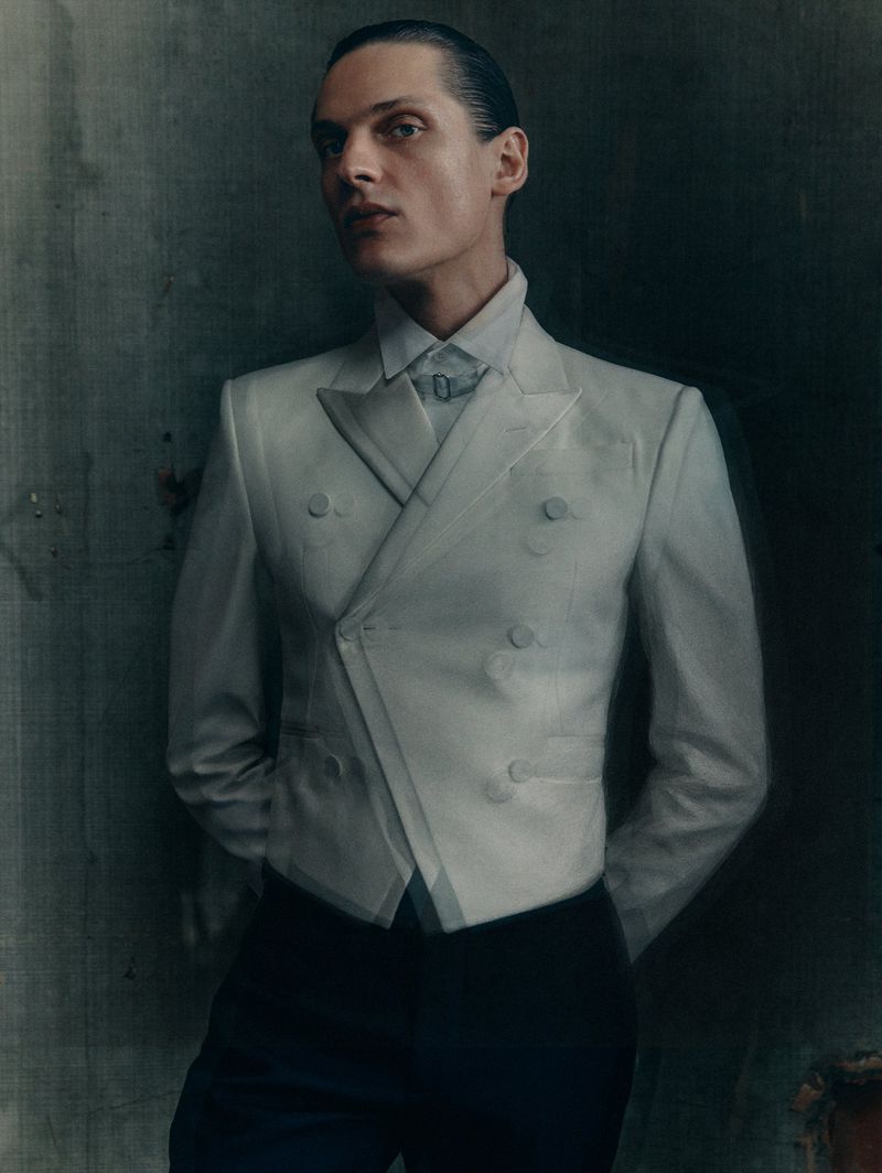 Portrait of a male fashion model in a pale grey blazer and dark trousers, standing indoors against a mottled grey wall in a studio setting.