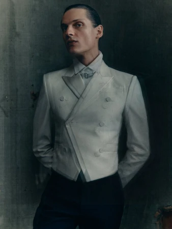 Portrait of a male fashion model in a pale grey blazer and dark trousers, standing indoors against a mottled grey wall in a studio setting.