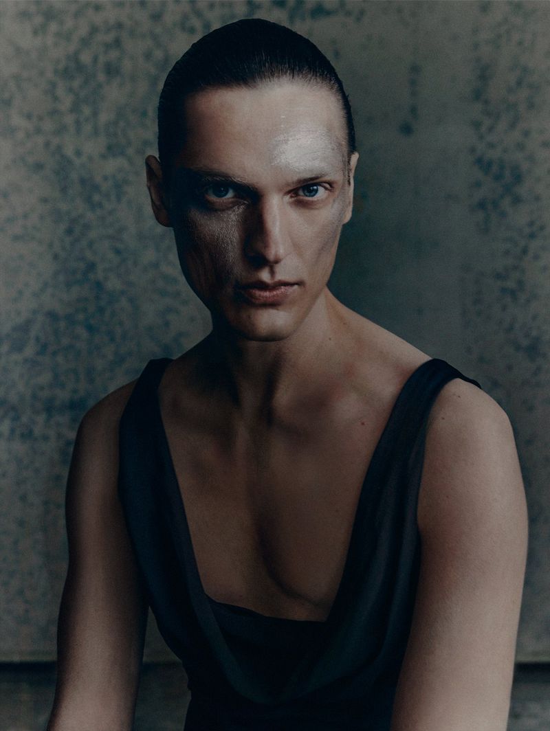 Close-up studio portrait of a person with slicked-back dark hair, light skin, and a neutral expression, wearing a black sleeveless top.