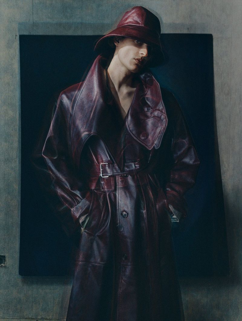 Fashion portrait of a person wearing a red leather trench coat and bucket hat, standing against a muted blue and grey backdrop.