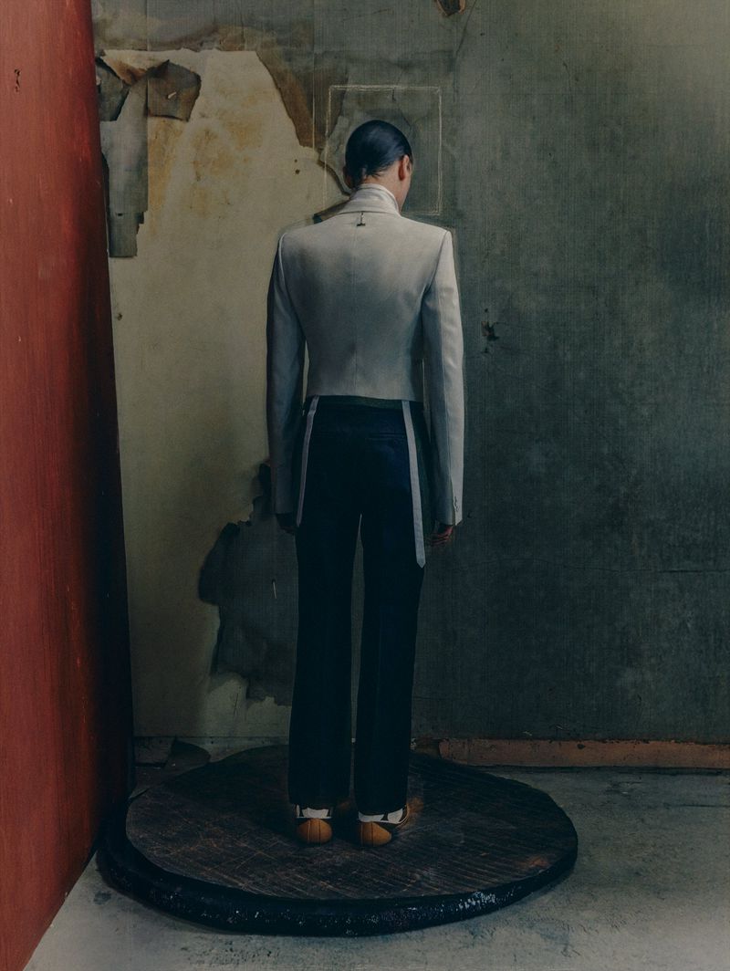 Back view of a person in a grey suit with dark pants, standing on a dark round wooden platform against an aged, weathered wall.