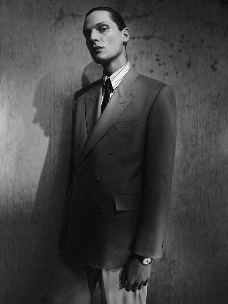 Black and white portrait of a man in a double-breasted suit with tie standing in front of a weathered wall with shadow.