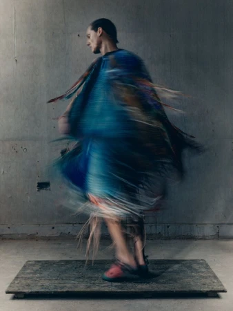 Model Valentin Caron spins on a low wooden platform wearing a blue fringe garment captured with dynamic motion blur against a concrete wall.