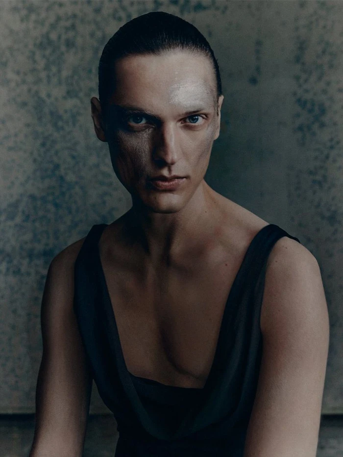 Portrait of a man wearing a dark sleeveless top with textured makeup on his face against a mottled wall background.