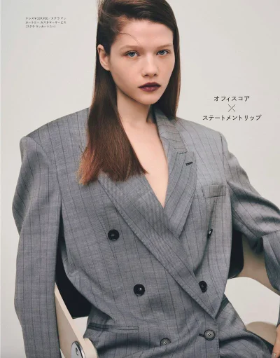 Model Eugene Dranitsa wearing an oversized grey pinstripe double-breasted blazer with bold dark lipstick against a pale studio backdrop.