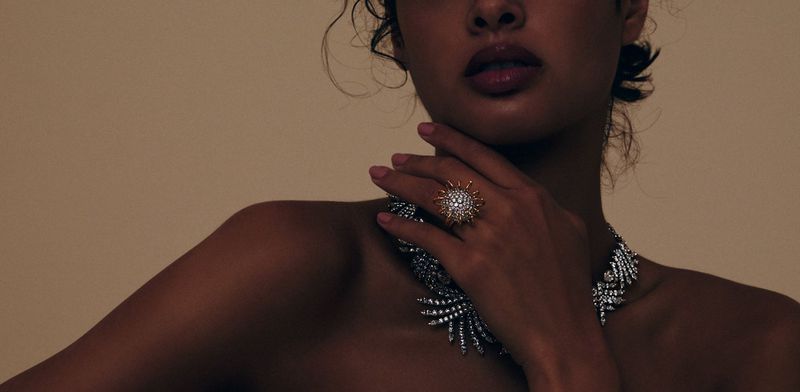 Close-up of a woman with dark skin wearing a glittering ring and necklace, posing in a photo studio with a neutral tan backdrop.