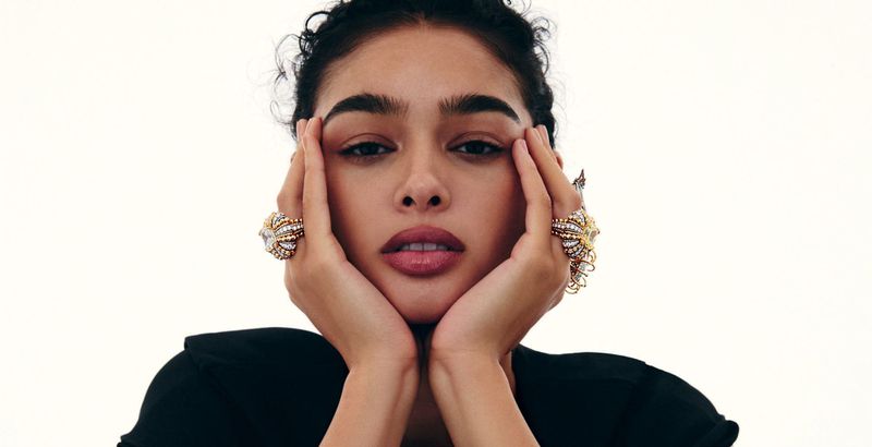 Close-up beauty portrait of a woman resting her face in her hands, wearing large ornate gold and diamond rings against a white backdrop.