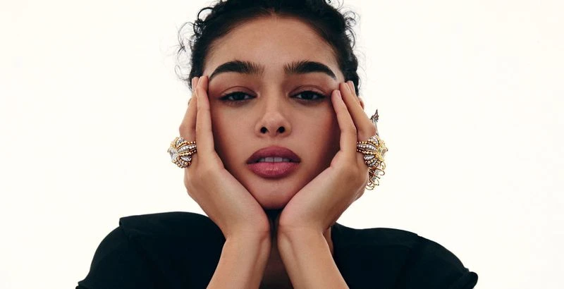 Close-up beauty portrait of a woman resting her face in her hands, wearing large ornate gold and diamond rings against a white backdrop.