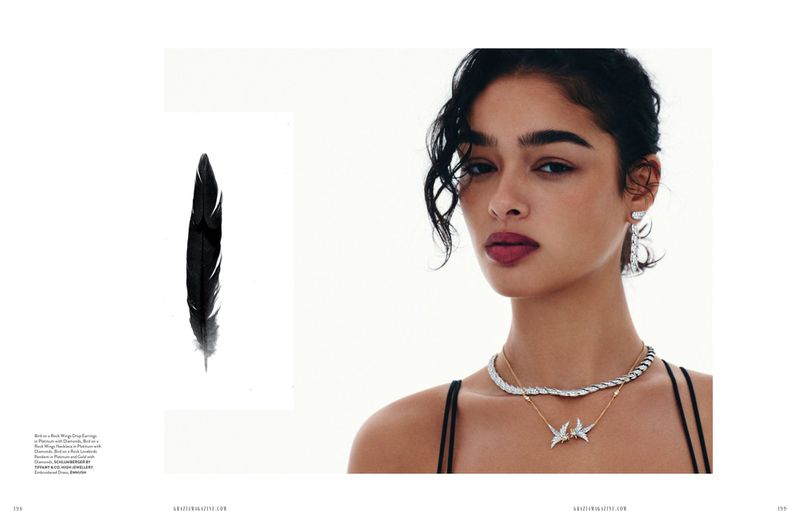 Close-up of a woman wearing jewelry, including earrings and a layered necklace, alongside a feather, against a white background.