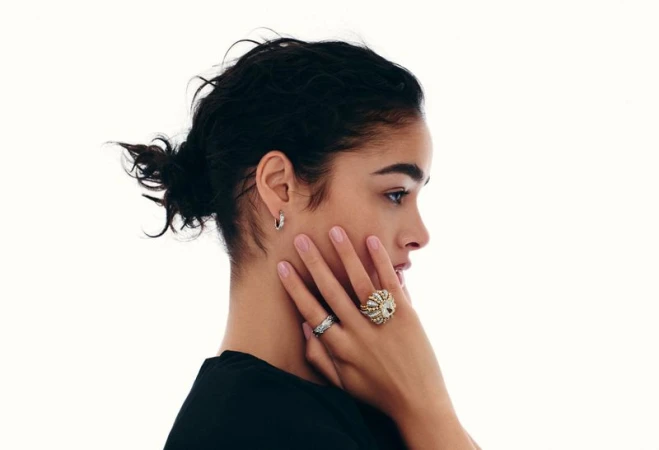 Profile portrait of a woman touching her face while showcasing large diamond cocktail rings and a small hoop earring.