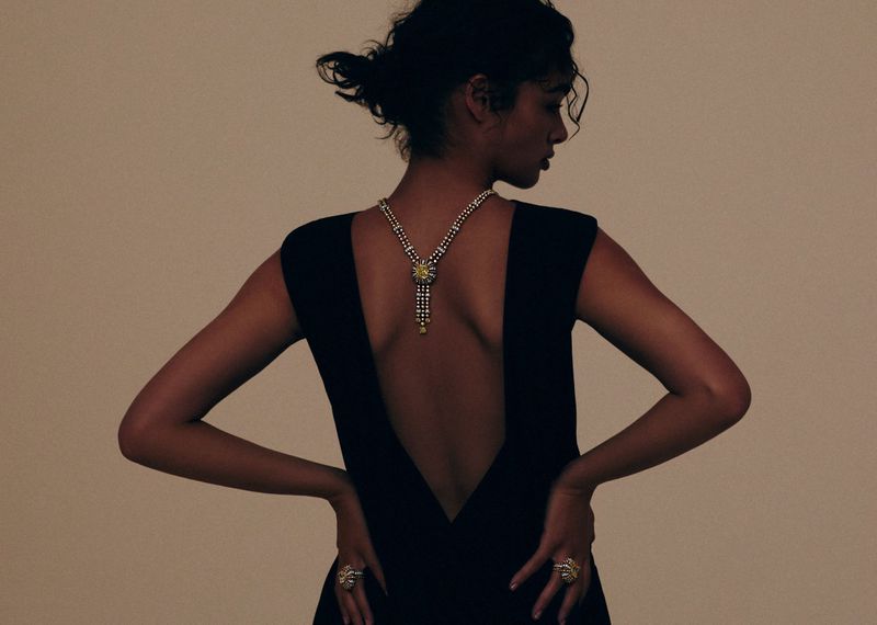 Woman in a black dress with an open back faces away from the camera, showing off a pearl necklace and a ring, against a neutral backdrop.