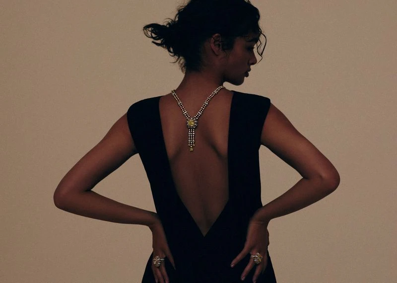 Woman in a black dress with an open back faces away from the camera, showing off a pearl necklace and a ring, against a neutral backdrop.