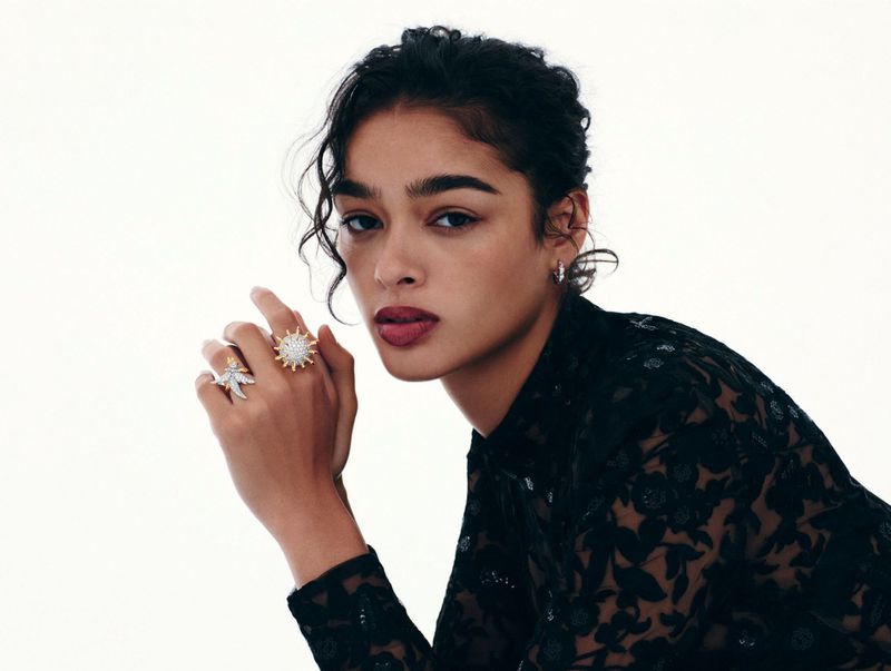 Portrait of a South Asian woman with a serious expression, wearing ornate rings and a floral lace top against a bright white backdrop.