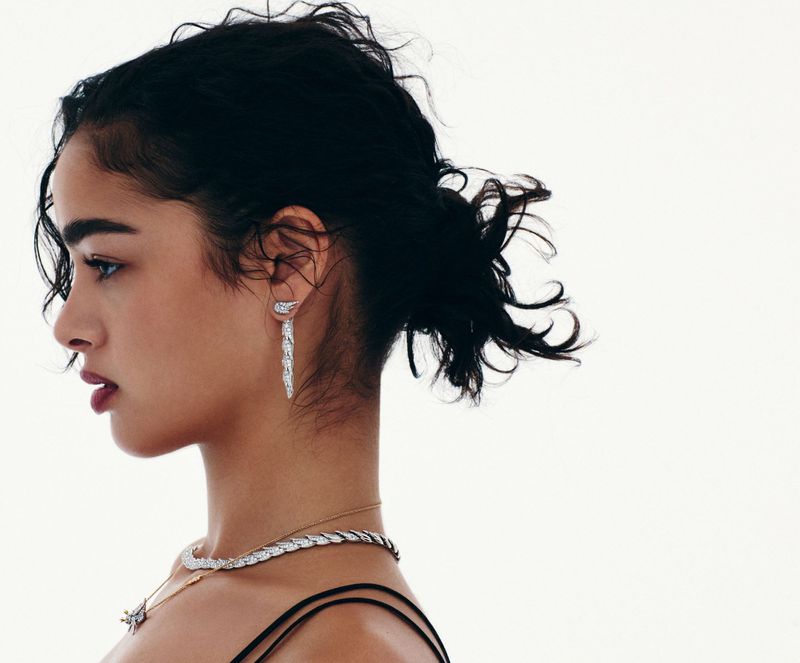 Profile view of an adult woman with dark curly hair wearing elegant jewelry, including a necklace and dangling earrings against a white background.