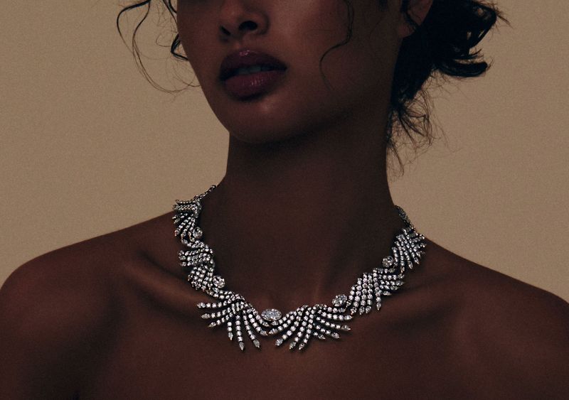 Adult woman with fair skin and dark curly hair poses indoors wearing an ornate crystal necklace against a cream-colored backdrop.
