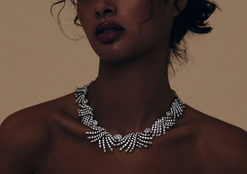 Adult woman with fair skin and dark curly hair poses indoors wearing an ornate crystal necklace against a cream-colored backdrop.