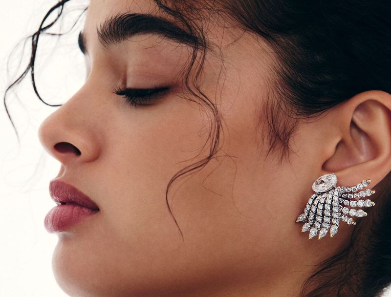Profile shot of a woman with dark hair, wearing large jeweled earrings, against a bright, neutral background. Beauty and fashion shot.