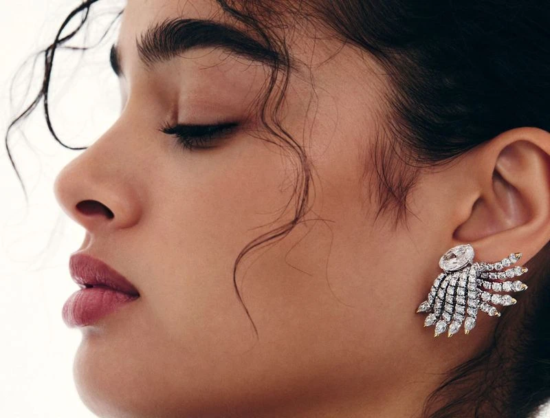 Profile shot of a woman with dark hair, wearing large jeweled earrings, against a bright, neutral background. Beauty and fashion shot.