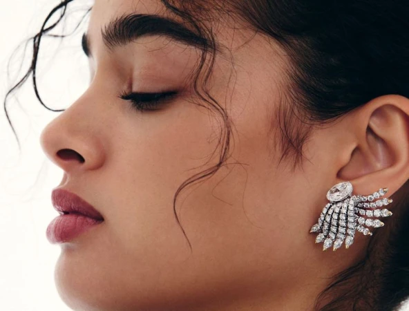 Profile shot of a woman with dark hair, wearing large jeweled earrings, against a bright, neutral background. Beauty and fashion shot.