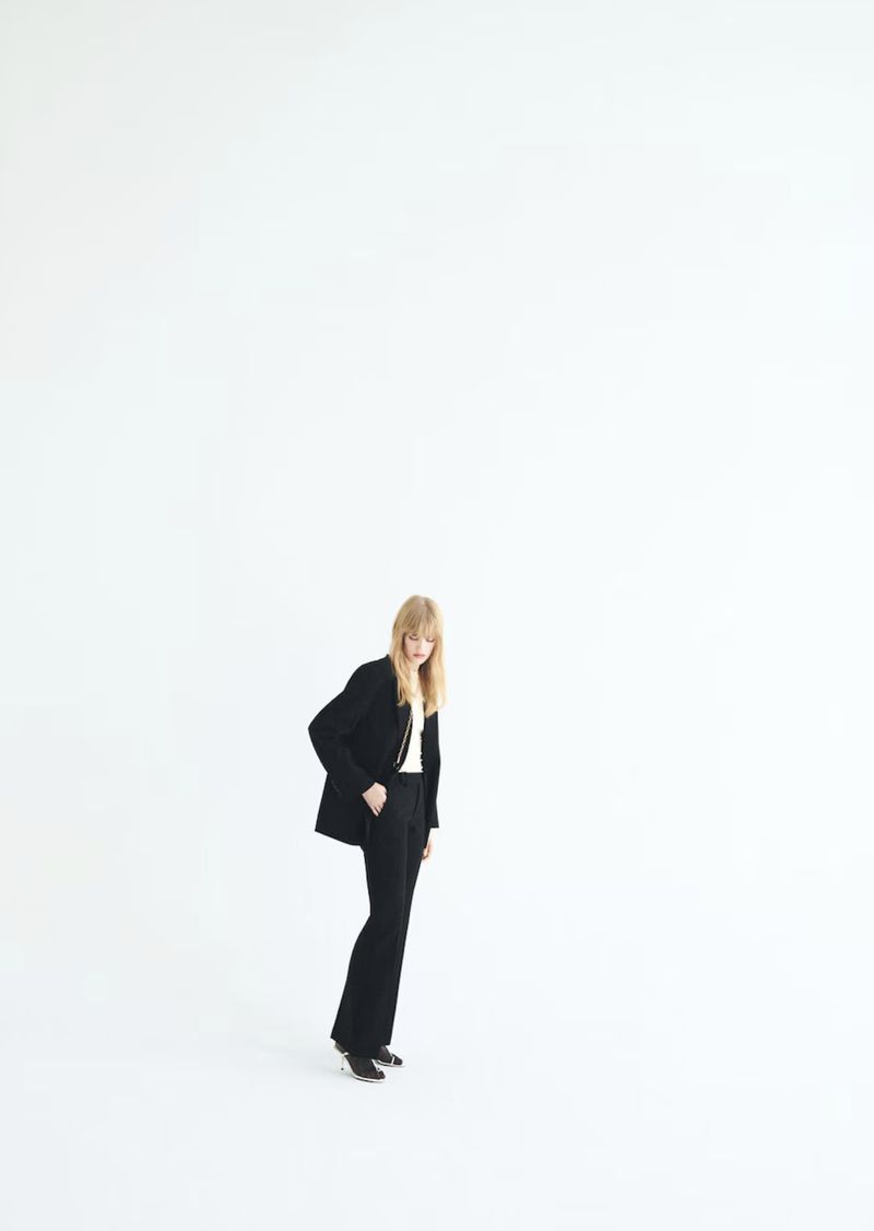 A woman in a black blazer and pants poses in a full shot against a bright white background in a fashion photography style.
