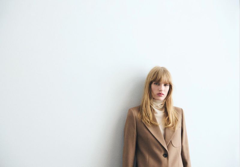 A blonde woman in a tan blazer and turtleneck poses against a white background in a fashion editorial shot with soft, natural light.