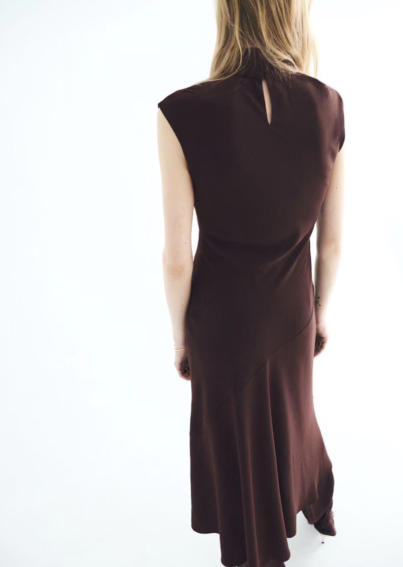 A slender woman in a brown sleeveless dress with a high neckline is seen from the back in a minimalist studio setting.