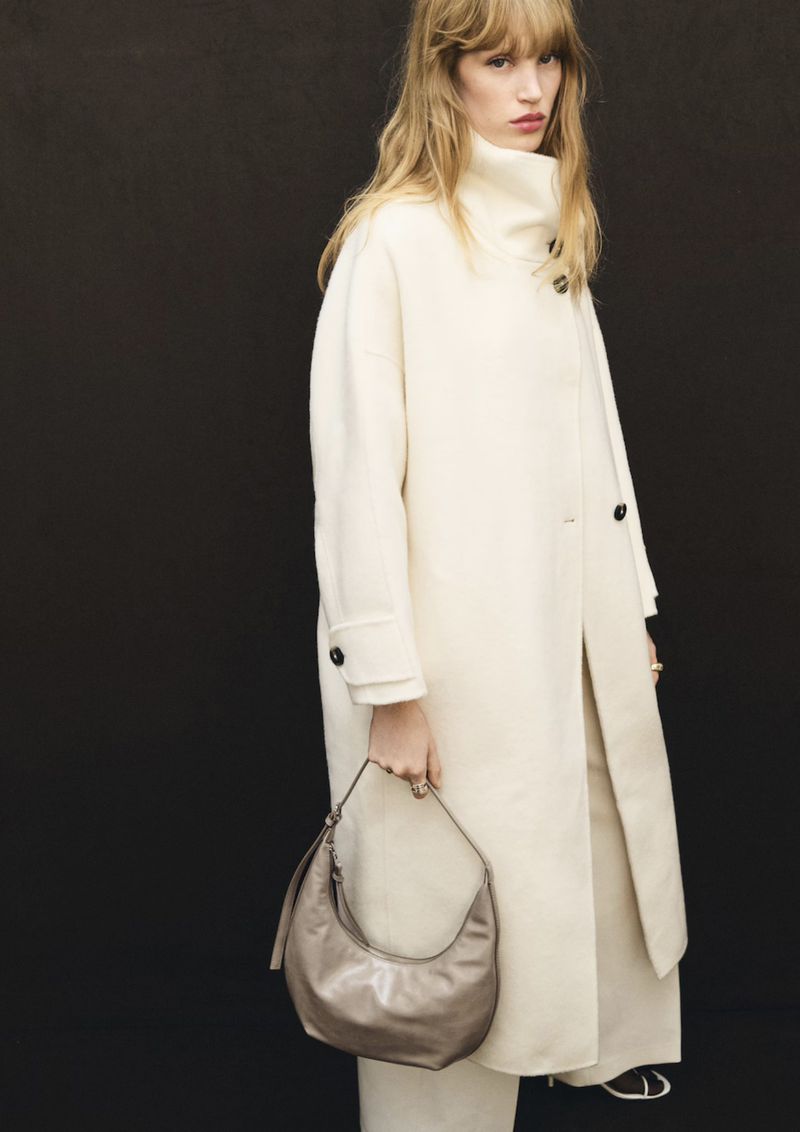 Model Jacqui Hooper wears an off-white high-neck coat while holding a taupe shoulder bag against a black seamless backdrop.