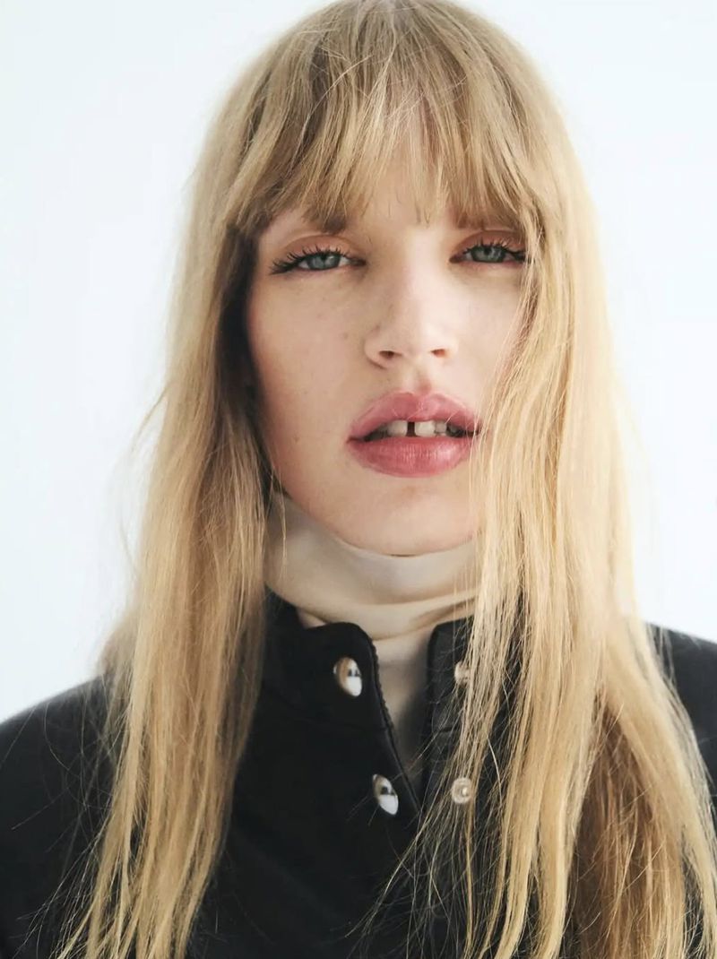 Close-up portrait of a blonde model with bangs wearing a black jacket and cream turtleneck against a plain background.