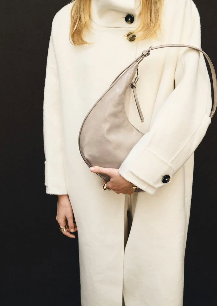 Model wearing an off-white buttoned coat holding a taupe leather curved shoulder bag against a solid black background.
