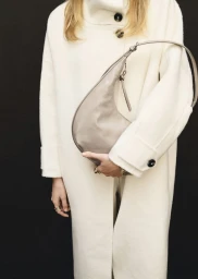 Model wearing an off-white buttoned coat holding a taupe leather curved shoulder bag against a solid black background.