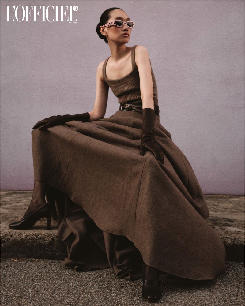 Fashion editorial shot of a woman in a brown dress, gloves, and sunglasses, seated on a stone surface with a muted purple background.