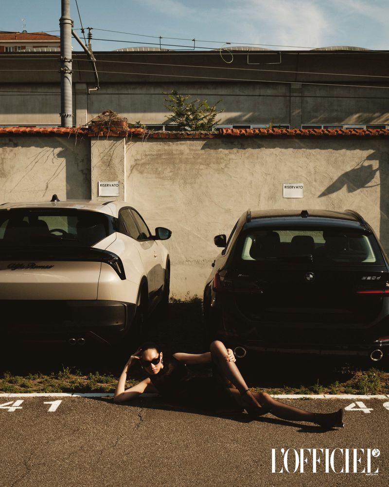 Fashion editorial shot outdoors: woman in sunglasses poses on asphalt between parked cars, magazine logo visible; sunny, daytime.