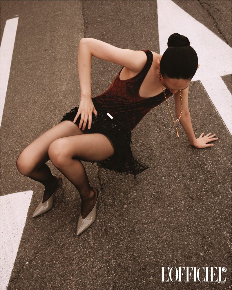 Woman in dress and heels poses on a city street with asphalt and painted white markings in a fashion editorial shot with neutral coloring.