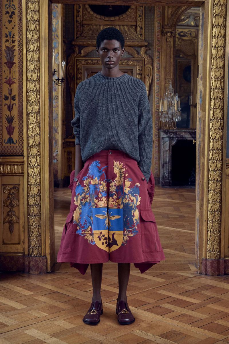 Fashion shot of an African-American man wearing a grey sweater and ornate burgundy shorts, indoors in a space with detailed mouldings.