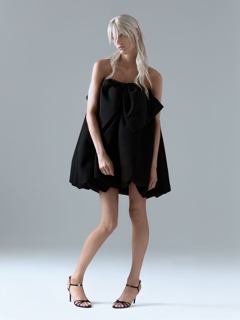 Woman in black dress posing in a studio. The black dress has a bow design. She is also wearing heeled sandals. Studio lighting.