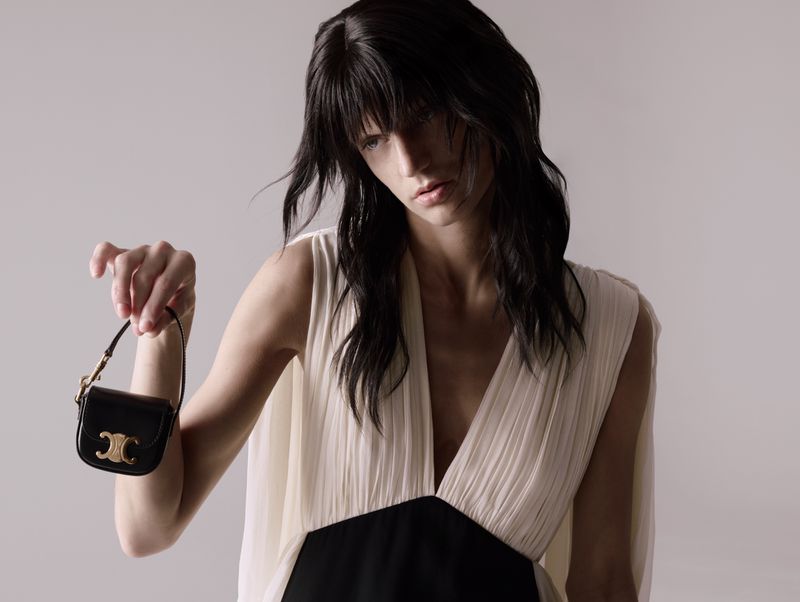 Brunette woman poses with a black leather mini-handbag with gold hardware in a fashion studio, modeling a sleeveless white top.