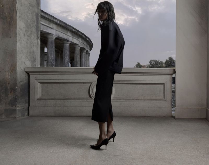 Elegant woman in a long black dress and heels posing against an old building with columns creates a timeless fashion image.
