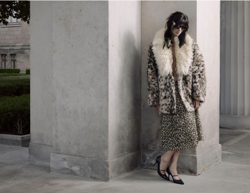 A woman in a cheetah print coat and skirt stands outside a building with columns, modeling fashion in an outdoor setting.
