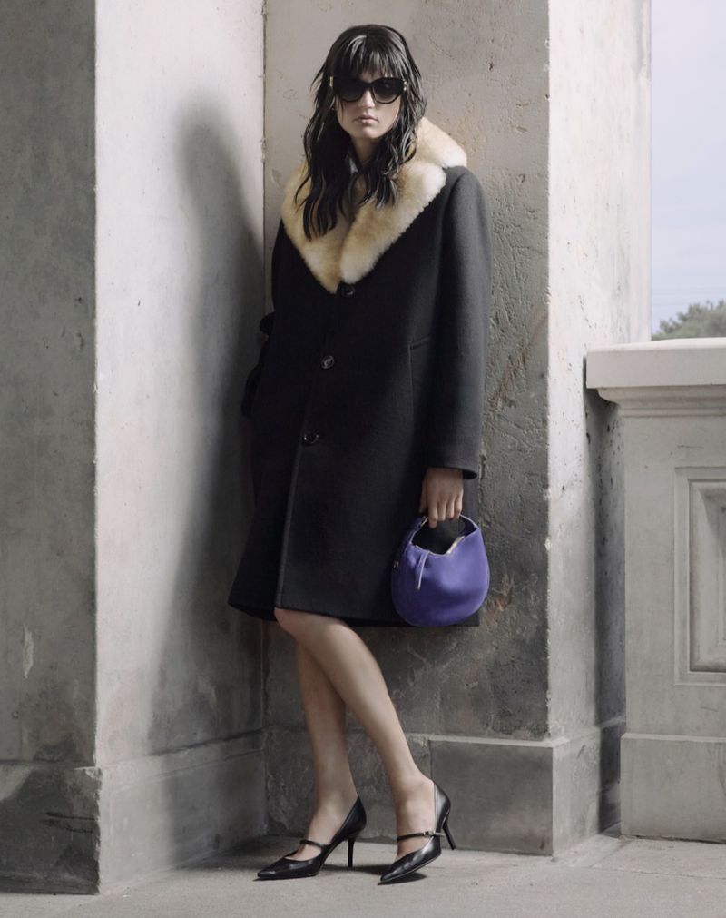 A woman in a black coat with a fur collar, sunglasses, and heels stands next to a corner wall holding a purple handbag.