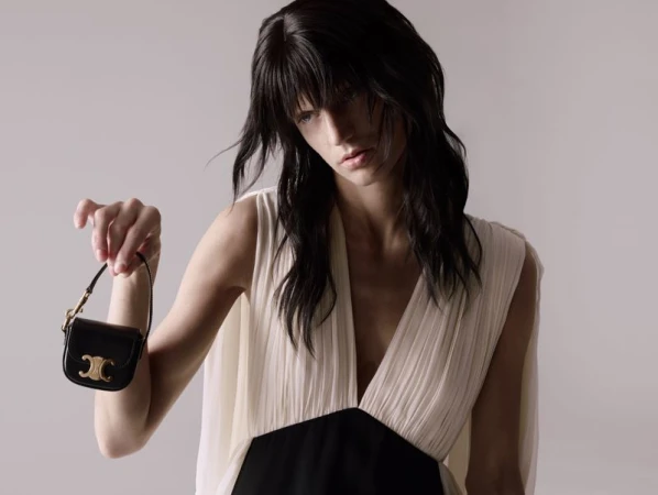 Model with shaggy dark hair holding a miniature black Celine handbag with gold hardware in front of a neutral grey backdrop.
