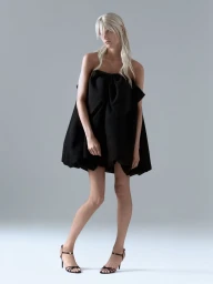 Model in a black strapless bow babydoll mini dress and black strappy heels against a neutral studio backdrop.