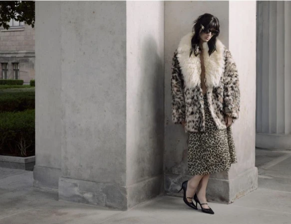 Fashion model in a plush leopard print coat, matching skirt, sunglasses, and black pointed heels standing by stone columns.