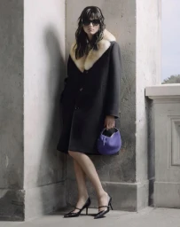 Model wearing a black fur-collared coat, sunglasses, pointed black heels, and holding a purple bag outdoors.